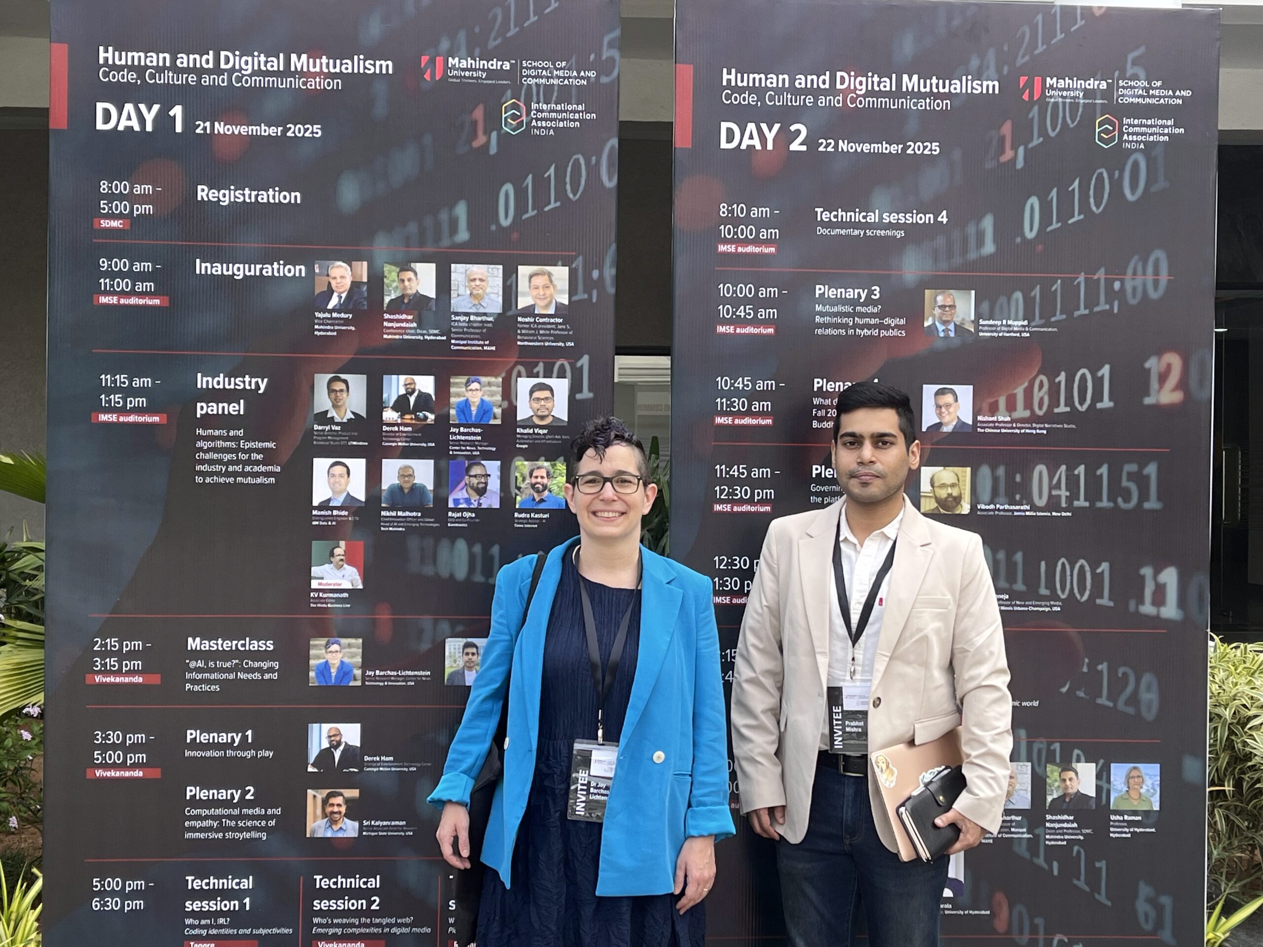A white person and Indian man standing side by side in front of the event poster.
