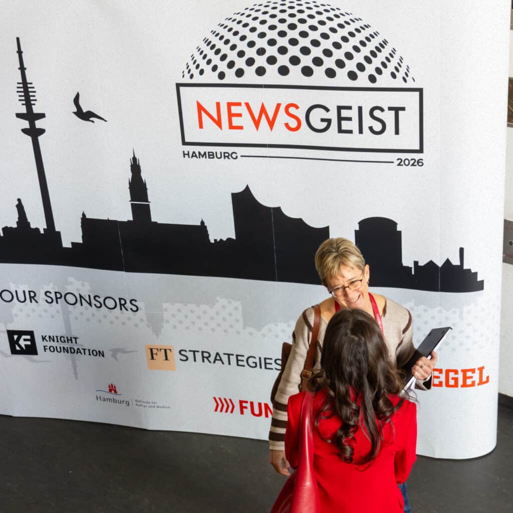 Two woman talking in front of banner featuring Newsgeist logo and sponsor logos.